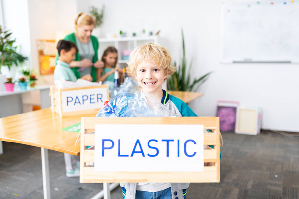 Χαρούμενο ξανθό αγόρι κρατώντας κουτί με πλαστικό μετά το μάθημα Οικολογία - Φωτογραφία, εικόνα