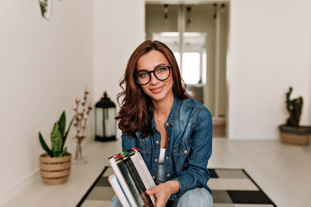 Feliz encantadora bela senhora vestindo óculos trabalhando com livros olhando para a câmera no fundo do apartamento moderno
  - Foto, Imagem