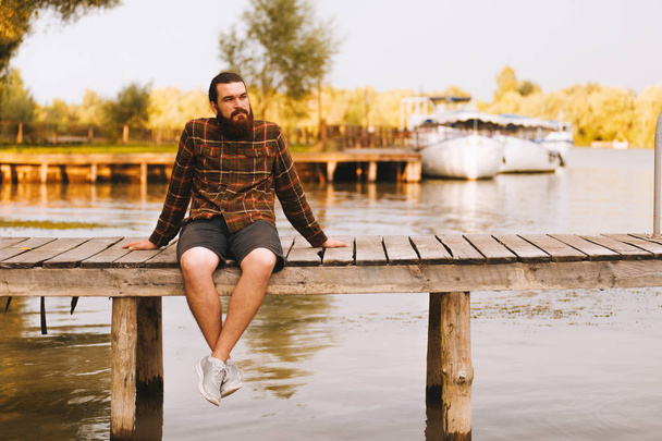 Young bearded man sitting on a berth upon a river. Green sunny b - Zdjęcie, obraz