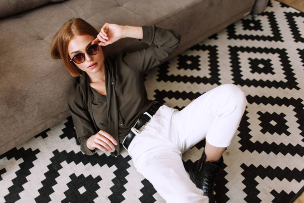 Beautiful sweet girl lying on the sofa. Golden stylish model with short red hair, plump lips, sunglasses, casual clothing, costume, outfit, overalls. Model test, advertising glasses, clothes - Fotografie, Obrázek