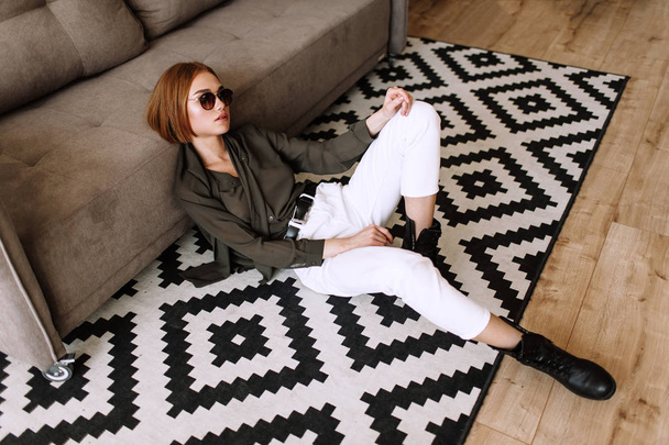 Beautiful sweet girl lying on the sofa. Golden stylish model with short red hair, plump lips, sunglasses, casual clothing, costume, outfit, overalls. Model test, advertising glasses, clothes - 写真・画像