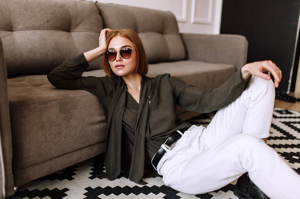 Beautiful sweet girl lying on the sofa. Golden stylish model with short red hair, plump lips, sunglasses, casual clothing, costume, outfit, overalls. Model test, advertising glasses, clothes - Foto, afbeelding