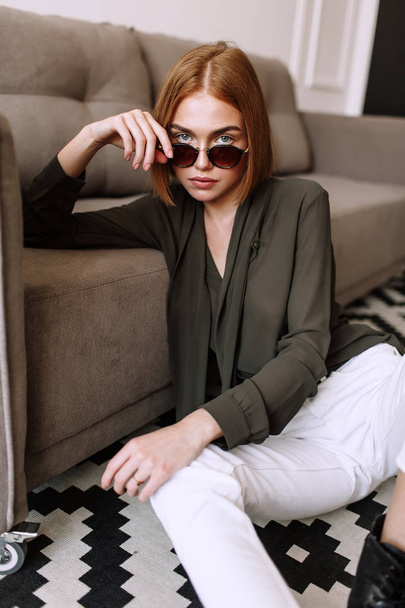 Beautiful sweet girl lying on the sofa. Golden stylish model with short red hair, plump lips, sunglasses, casual clothing, costume, outfit, overalls. Model test, advertising glasses, clothes - Фото, изображение