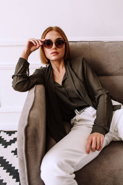Beautiful sweet girl lying on the sofa. Golden stylish model with short red hair, plump lips, sunglasses, casual clothing, costume, outfit, overalls. Model test, advertising glasses, clothes - Φωτογραφία, εικόνα