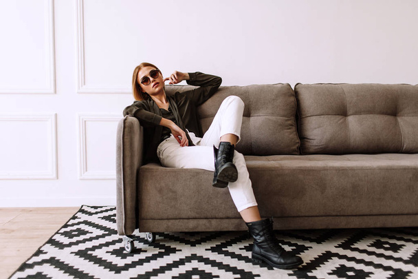 Beautiful sweet girl lying on the sofa. Golden stylish model with short red hair, plump lips, sunglasses, casual clothing, costume, outfit, overalls. Model test, advertising glasses, clothes - Photo, image