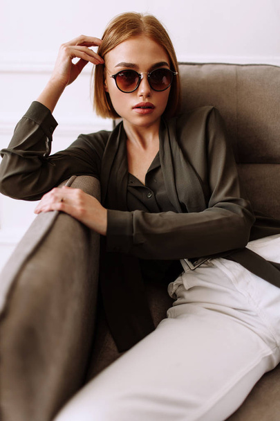 Beautiful sweet girl lying on the sofa. Golden stylish model with short red hair, plump lips, sunglasses, casual clothing, costume, outfit, overalls. Model test, advertising glasses, clothes - Foto, immagini