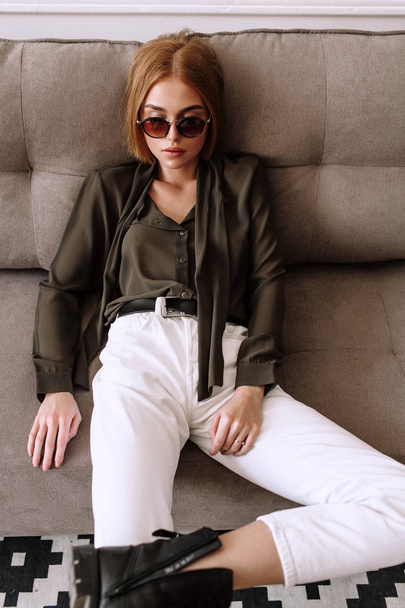 Beautiful sweet girl lying on the sofa. Golden stylish model with short red hair, plump lips, sunglasses, casual clothing, costume, outfit, overalls. Model test, advertising glasses, clothes - Foto, Imagen