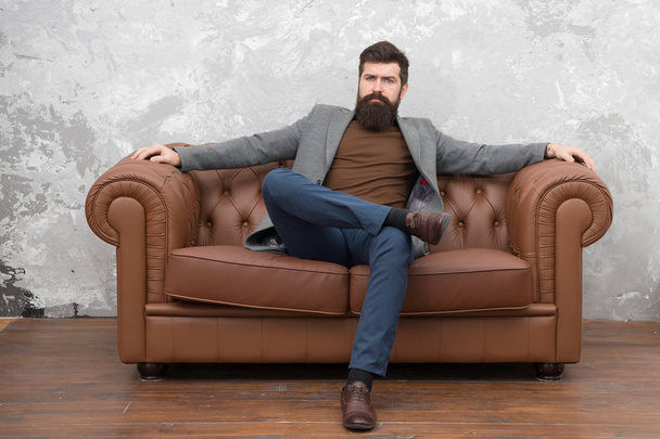 Bearded man with confident face sit leather couch. Loft interior apartment. Businessman realtor work. Furniture shop. Hipster realtor loft style apartment. Realtor and rental service. Rent apartment - Foto, afbeelding