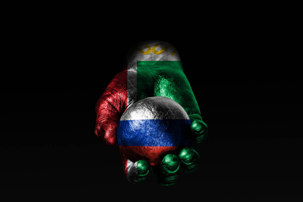 Una mano con una bandera chechena dibujada sostiene una pelota con una bandera rusa dibujada, un signo de influencia, presión o conservación y protección
. - Foto, Imagen