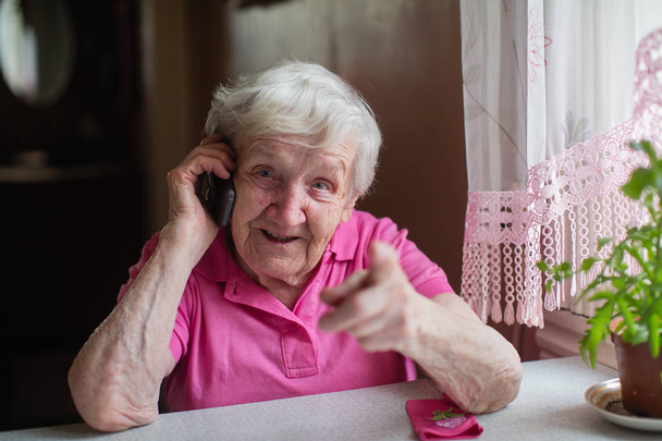 Vanha nainen puhuu puhelimessa ja istuu pöydässä kotonaan.
. - Valokuva, kuva
