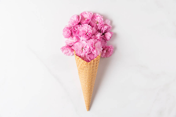 Ice cream cone with pink cherry blossoming flowers bouquet on white marble background. Minimal spring concept - Zdjęcie, obraz