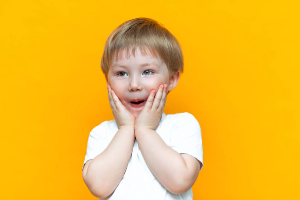cheerful baby boy three years old in white t-shirt stands on yellow background, open his mouth surprised, blonde hair mixed race asian - Foto, immagini