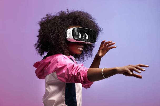 Mod young brown-haired curly girl dressed in the pink sports jacket uses the virtual reality glasses in the studio on neon background - Photo, Image