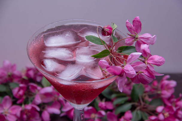 Słodki koktajl z lodem przelewa się do kieliszka do Martini, krople zimnej wody spływały ściany szkła, szkło i stół są ozdobione gałęziami kwitnących drzew z różowymi kwiatami. - Zdjęcie, obraz