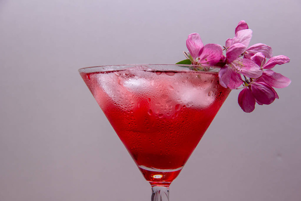Una bebida alcohólica dulce con hielo se vierte en una copa de martini, el vidrio está decorado con una rama de un árbol de flores de primavera con flores de color rosa
. - Foto, Imagen