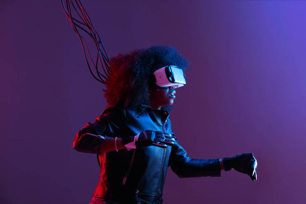 Mod curly dark haired girl dressed in black leather jacket and gloves uses the virtual reality glasses on her head in the dark studio with neon light - Photo, image