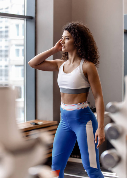 Young good fit curly girl dressed in sports clothes is standing next to the sports equipment in the modern gym by the window - Zdjęcie, obraz