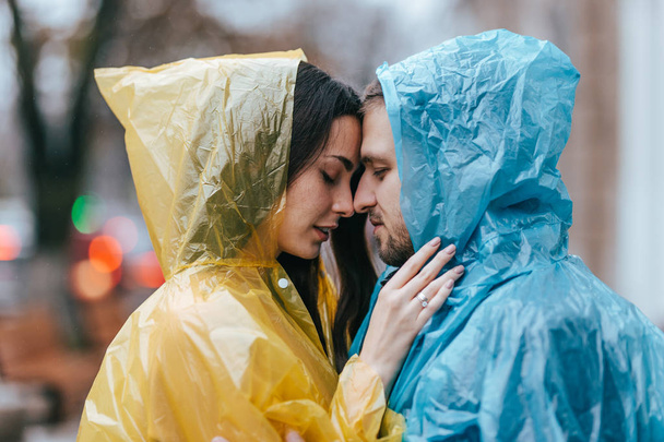 Loving guy and his girlfriend in the raincoats stand face to face on the street in the rain - 写真・画像