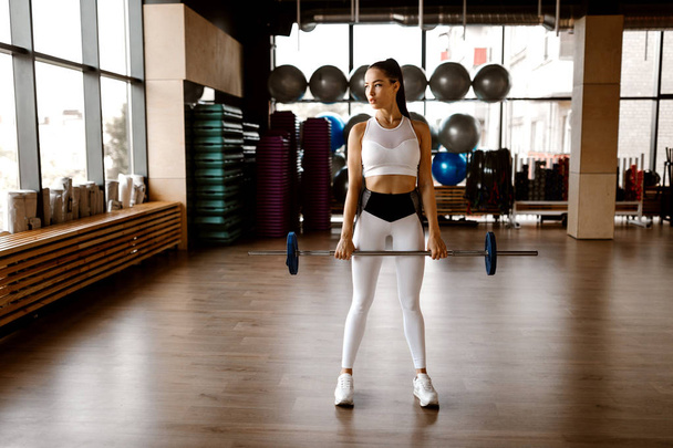 Athletic girl  dressed in white sports top and tights holds barbell in the gym - Фото, зображення
