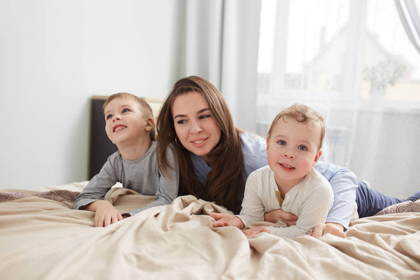 Happy family. Young mother dressed in light blue pajama lays with her two little sons on the bed with beige blanket in the bedroom with big window - Photo, Image