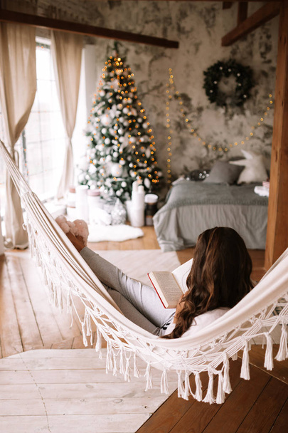 Beautiful dark-haired girl dressed in pants, sweater and warm slippers reads a book lying in a hammock in a cozy decorated  room with a New Year tree - Fotoğraf, Görsel