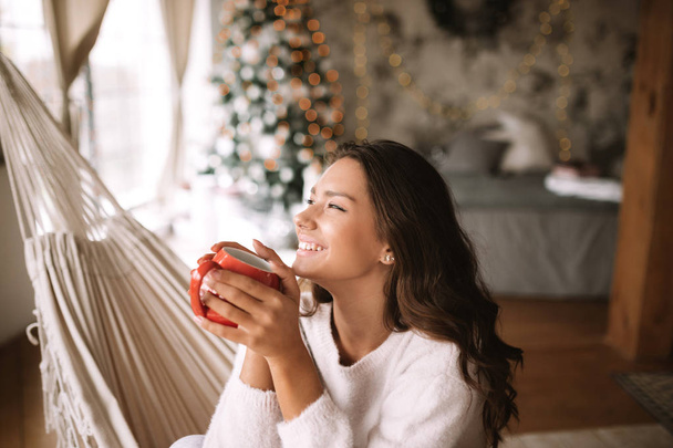 Smiling dark-haired girl dressed in beige sweater and pants holds a red cup sitting in a hammock in a cozy decorated room with a New Year tree - Φωτογραφία, εικόνα