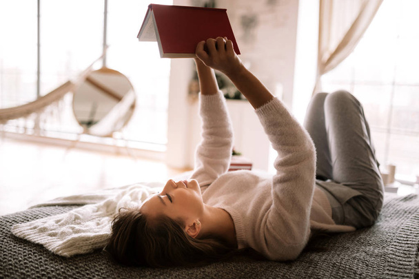 Charming girl dressed in white sweater and pants reads a book liying on the bed with gray blanket, white pillows and a New Year gift in a cory room - Photo, image
