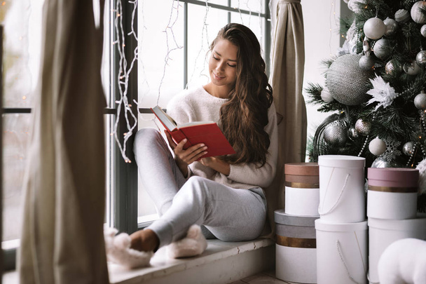 Nice dark-haired girl dressed in pants, sweater and warm slippers reads a book sitting on the windowsill of a panoramic window in the room next to the New Year tree, gifts and candles - Photo, Image