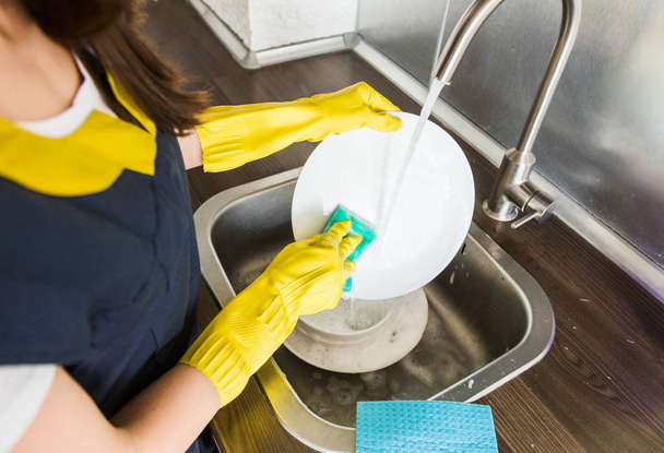 Een jonge vrouw met gele handschoenen wast de vaat met een spons in de gootsteen. Professionele schoonmaakdienst. - Foto, afbeelding