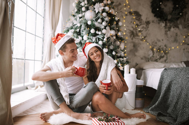 Happy guy and girl in white t-shirts and Santa Claus hats sit with red cups on the floor in front of the window next to the New Year tree, gifts and candles - Foto, afbeelding