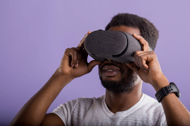 Handsome young African man in VR headset standing against purple background - Foto, Imagen
