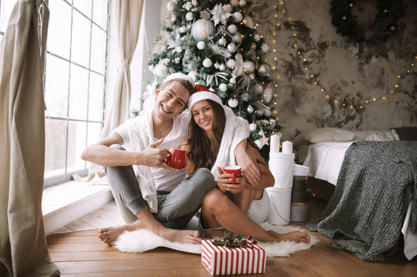 Happy guy and girl in white t-shirts and Santa Claus hats sit with red cups on the floor in front of the window next to the New Year tree, gifts and candles - Φωτογραφία, εικόνα