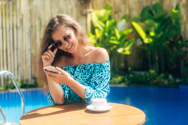 Beautiful model in sunglasses sits outdoors at table with cup of tea or coffee and uses phone - Foto, Bild