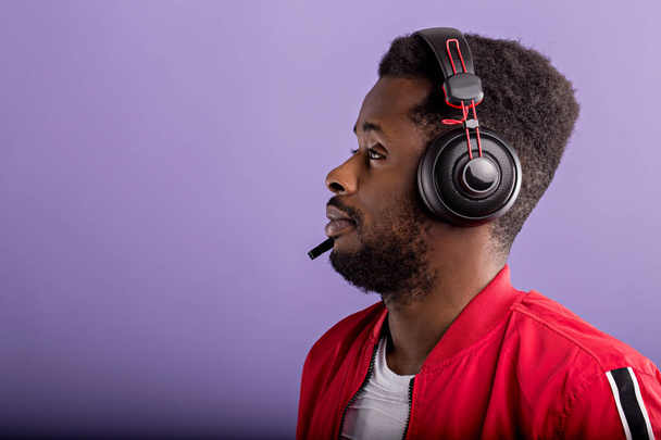 Side portrait of a beautiful afroamerican men in a red sports jacket and headphones on purple background with copy space - Φωτογραφία, εικόνα