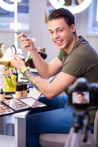 Joyful short-haired man sitting in studio lights and applying makeup - Fotografie, Obrázek