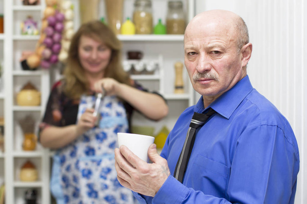 Пожилой мужчина с чашкой на фоне своей жены
 - Фото, изображение