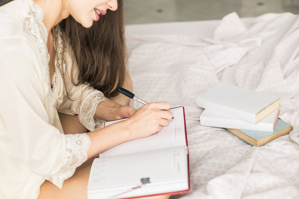 Attractive young girl in a nightgown is sitting in bed and writing into a notebook. - Foto, Imagem