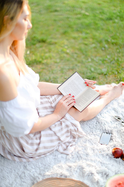 młoda blondynka biały kobieta czytanie książki i siedzi na kratę w pobliżu owoce w park. - Zdjęcie, obraz