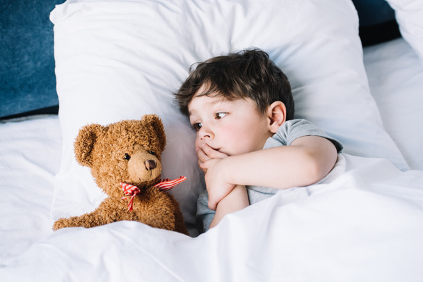 lindo niño acostado en la almohada blanca en la cama y mirando osito de peluche
  - Foto, Imagen