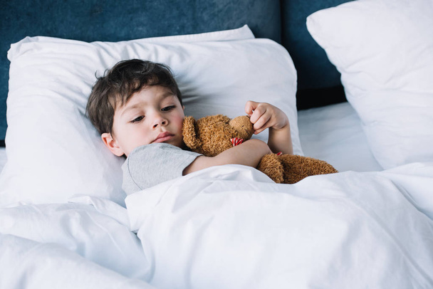 χαριτωμένο παιδί ξαπλωμένο σε λευκό μαξιλάρι στο κρεβάτι και κρατώντας αρκουδάκι  - Φωτογραφία, εικόνα
