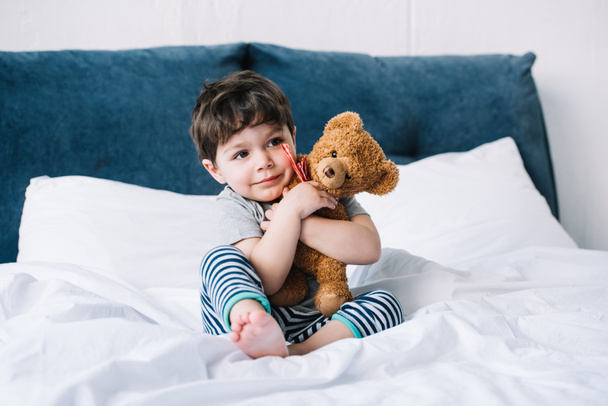 niño feliz con los pies descalzos sentado en la cama y abrazando oso de peluche
  - Foto, imagen