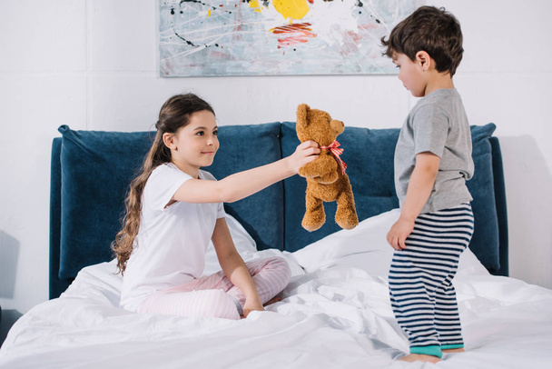 happy kid smiling while holding teddy bear near toddler brother in bedroom  - Photo, Image
