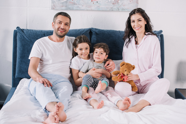 cheerful family with barefoot sitting on bed and smiling while looking at camera  - Zdjęcie, obraz