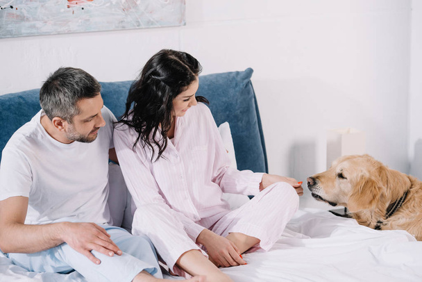 happy woman touching cute golden retriever while sitting on bed with man  - Fotó, kép