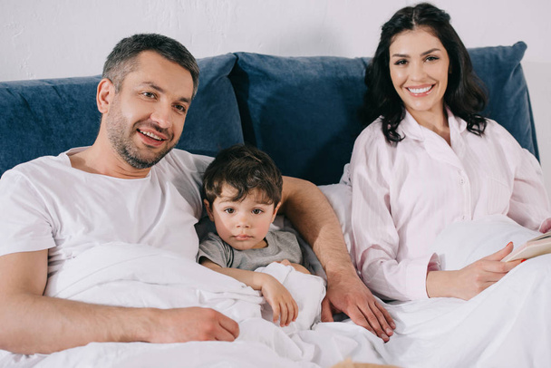 happy father smiling near toddler son and attractive wife in bedroom  - Фото, изображение