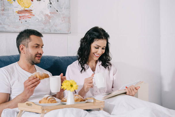 happy woman reading book near husband holding croissant in bed  - Φωτογραφία, εικόνα