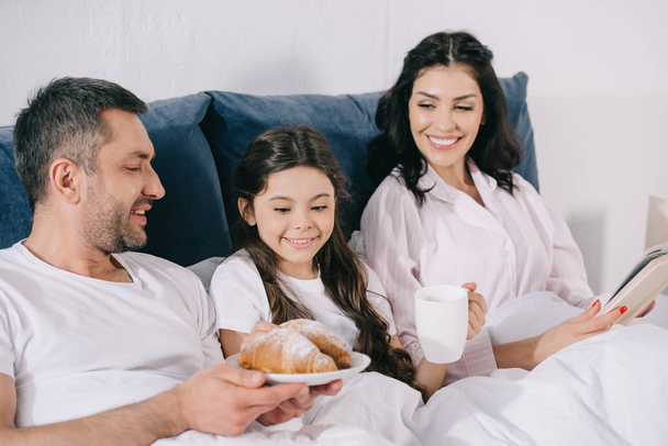cheerful woman holding book near daughter with cup and husband with croissants  - Foto, immagini