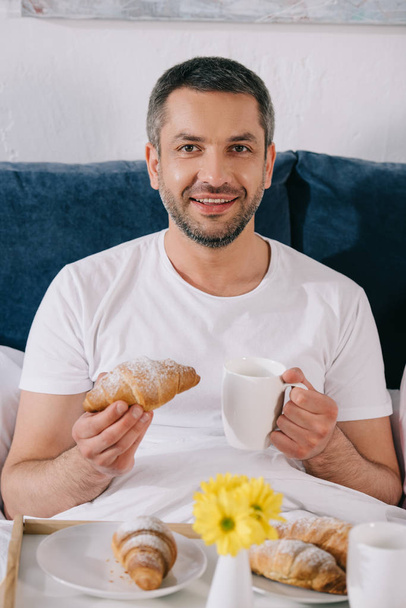 εύθυμος άνθρωπος κρατώντας γλυκό κρουασάν και φλιτζάνι καφέ στο κρεβάτι  - Φωτογραφία, εικόνα