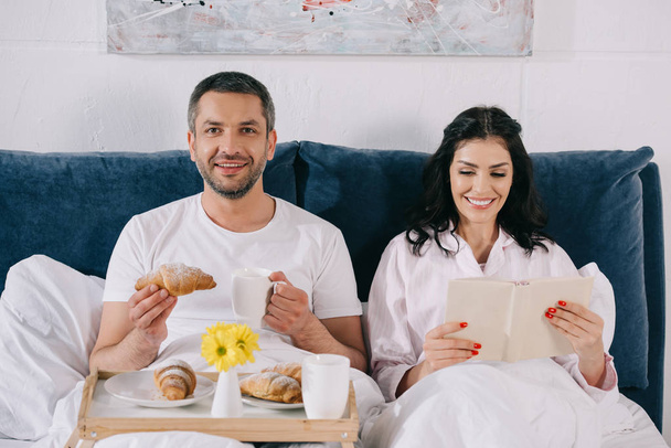 happy woman reading book near cheerful husband holding croissant and cup in bed  - Foto, afbeelding