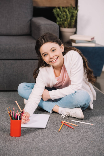 smiling kid looking at camera while holding pencil near paper in living room  - Foto, immagini